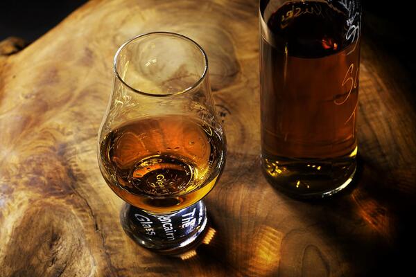 Ein halb gefülltes Glas mit bernsteinfarbenem Whiskey und eine ebenfalls damit gefüllte Flasche stehen auf einem natürlich gemusterten Holztisch.