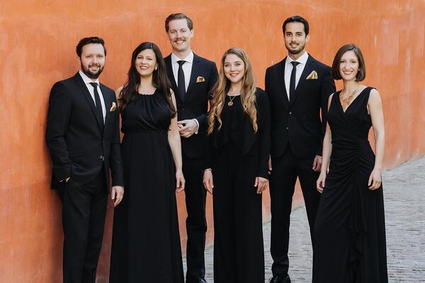 Bild vergrößern: Die Musikerinnen und Musiker des Vokalensembles Stimmgold stehen in festlicher schwarzer Abendgarderobe vor einer roten Wand.