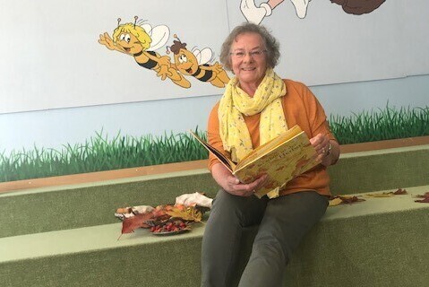 Die Autorin Ingeborg Neger sitzt mit einem aufgeschlagenen Bilderbuch in der Hand vor einer mit Zeichentrickfiguren bemalten Wand. 