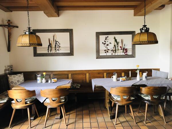 Zwei Bilder im Hintergrund an der Wand, davor stehen zwei Tische mit Stühlen aus Holz. Die Tische sind mit einer Decke und Vasen gedeckt.