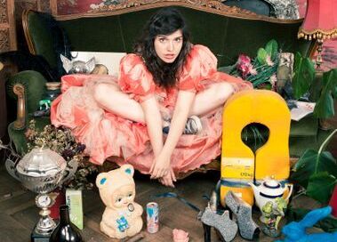 Die Sängerin Nele sitzt in einem rosa Rüschenkleid im Schneidersitz auf einer Bank. Vor ihr sind verschiedene Gegenstände, wie auf einem Flohmarkt aufgereiht. 