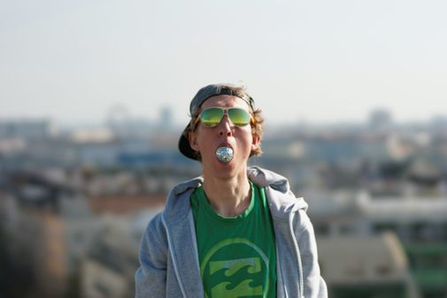 Der Musiker Tombo trägt eine verpsiegelte grüne Sonnenbrille, ein grünes T-Shirt und hat eine kleine Diskokugel im Mund. 