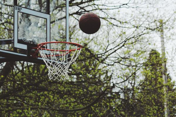 Ein Basketball fliegt in einen Basketballkorb. Im Hintergrund sind Bäume mit grünem Laub. 