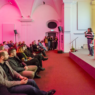Seitlicher Blick in den Innenraum der Spitalkirche. Links sitzt das Publikum und blickt auf den eine Frau in einem schwarz-weiß gestreiften Pullover, die auf einer Bühne im rechten Teil des Bildes steht.