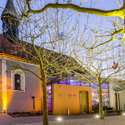 Die von außen beleuchtete Spitalkirche in der Dämmerung. In den kahlen Bäumen vor der Kirche hängen bunte Vögel aus Karton.