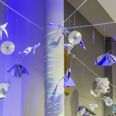Origami-Vögel und CDs hängen um eine mit Neon-Fäden umwickelte graue Säule.