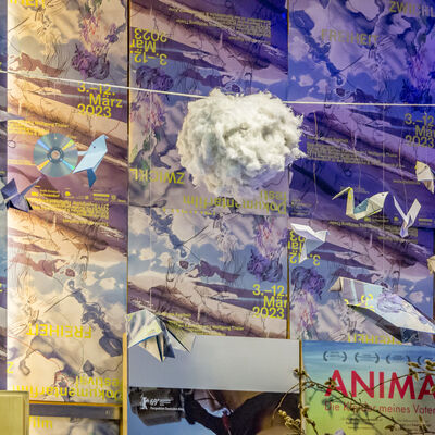 Vor einer mit Zwickl-Plakaten beklebten Wand hängen an einer Schnur eine Runde Wolke aus Watte, Origami-Vögel und CDs.
