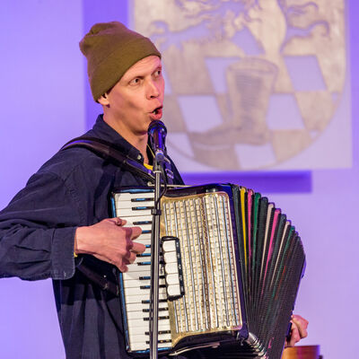 Der Musiker Maxi Pongratz singt in ein Mikrofon und spielt dabei Akkordeon.