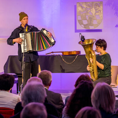 Der Musiker Maxi Pongratz steht auf der Bühne der Spitalkirche. Er singt in ein Mikrofon und spielt Akkordeon. Neben ihm sitzt die Musikerin Theresa Loibl, sie spielt Tuba.