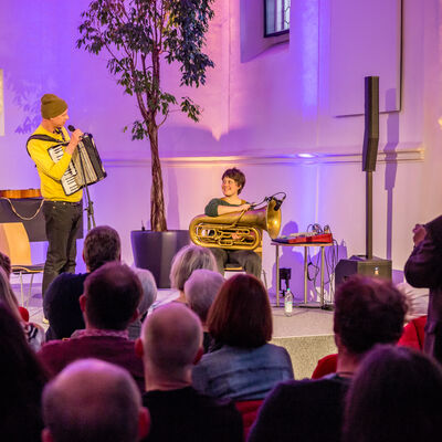 Der Musiker Maxi Pongatz hat ein Akkordeon umgehängt und spricht mir dem Publikum. Die Musikerin Theresa Loibl sitzt neben ihm. Sie hat eine Tuba auf dem Schoß.