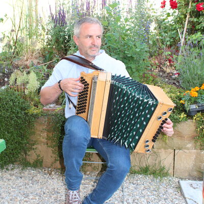 Bild vergrößern: Bierführung: "Das fünfte Element", der Musiker spielt mit seinem Harmonium, am Stuhl sitzend, auf. Hinter ihm blühen verschiedene Blumen.