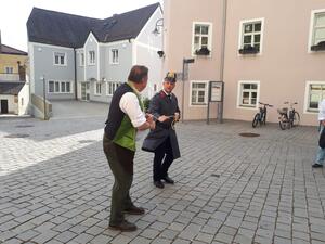 Bild vergrößern: Im Hintergrund befinden sich ein blaues und ein rosa Haus. Der Polizist in einer alten Uniform führt den Mann mit grünen Leibchen mit einem Strick ab.