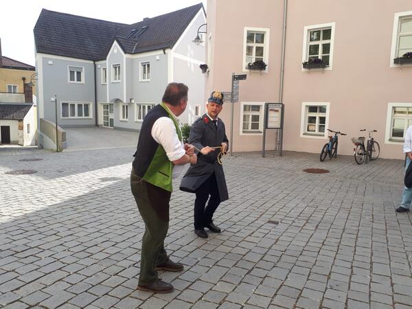 Im Hintergrund befinden sich ein blaues und ein rosa Haus. Der Polizist in einer alten Uniform führt den Mann mit grünen Leibchen mit einem Strick ab. 