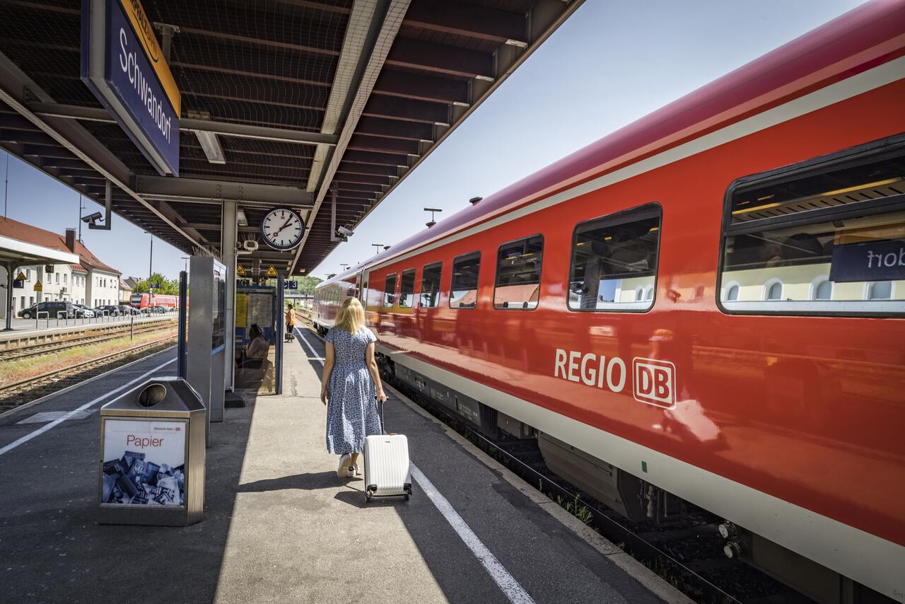 Bild vergr&ouml;&szlig;ern: Foto vom Schwandorfer Bahnhof. Man sieht einen roten Zug der Deutschen Bahn und eine Frau im blauen Kleid mit einem wei�en Koffer.