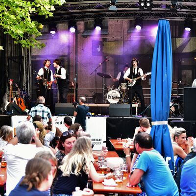 Bild vergrößern: Die Beatles-Coverband spielt auf der Bühne am Adolf-Kolping-Platz. Davor sitzt das Publikum an Biertischen.