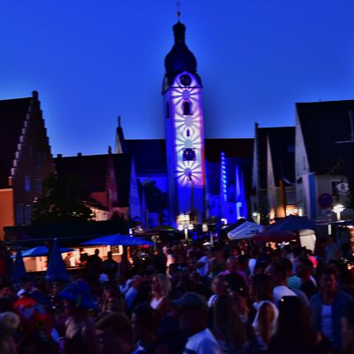 Bild vergrößern: Der mit einem blau-weißem Muster illuminierte Kirchturm St. Jakob beim Schwandorfer Bürgerfest. Davor sind umrisshaft viele Menschen auf dem Marktplatz zu sehen.
