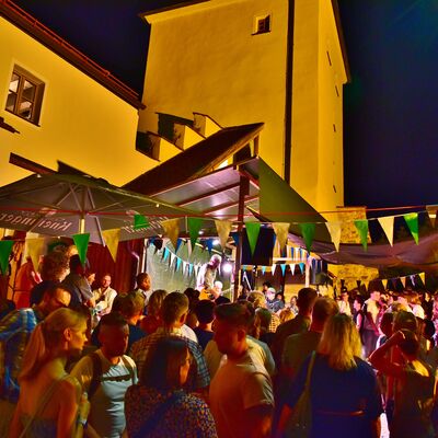 Bild vergrößern: Viele BesucherInnen versammeln sich vor der Bühne am Blasturm. Es ist dunkel, der Blasturm ist warm beleuchtet und der Festplatz mit grün-weißen Fähnchen geschmückt.