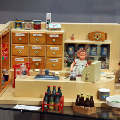 Bild vergrößern: Ein altes Puppenkaufladen.
Zu sehen ist ein kleiner Laden mit verschiedenen Lebensmitteln bei der Kassentheke.