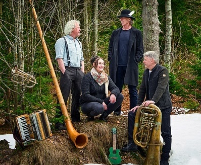 Bild vergrößern: Die Musiker Susi Raith, Edgar Feichtner, Franz Mosauer und Adrian Kreuzer versammeln sich um einen Strohballen vor einem schneebedeckten Wald. Um sie herum sind ihre Instrumente, ein Akkordeon, ein Alphorn, eine Ukulele und eine Tuba.