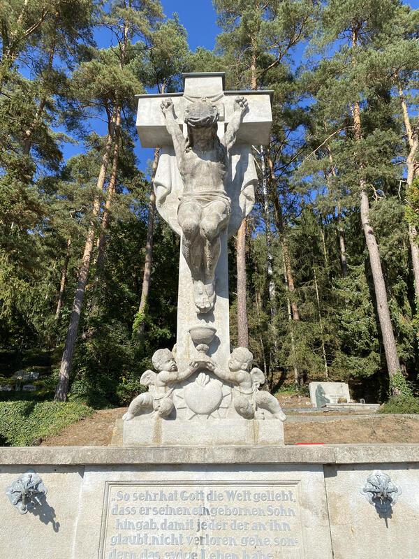 Bild vergr&ouml;&szlig;ern: Ein gro�es Kreuz aus Stein mit zwei Engeln, welche Jesus zu F��en liegen und einen Kelch zu ihm emporheben. Im Hintergrund stehen viele B�ume.