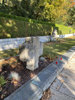 Bild vergrößern: Ein größeres Kreuz aus Stein steht für die Opfer des Kriegs am Friedhof, davor stehen noch zwei kleine rote Grablichter.