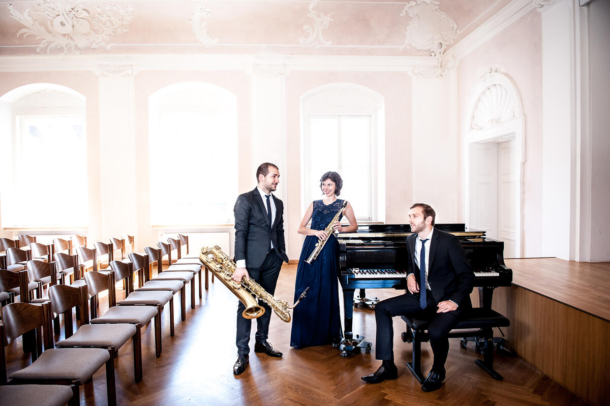 Ein Gruppenfoto der Band Trio Etoiles. Sie tragen Abendgarderobe, halten Blasinstrumente in der Hand und stehen neben einem Piano.