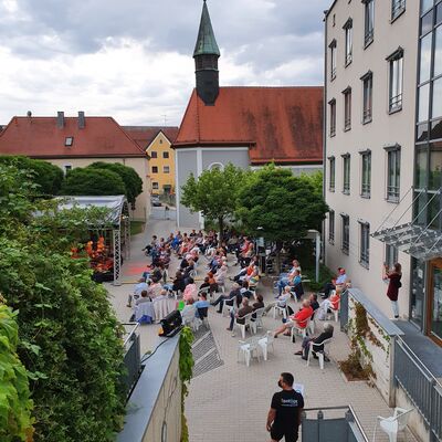 Bild vergrößern: Foto des Spitalgartens mit der Open-Air-Bühne. Zahlreiche Personen sitzen vor einer Bühne. Im Hintergrund ist die Spitalkirche zu sehen.