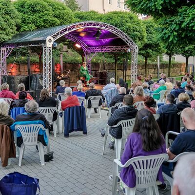 Bild vergrößern: Foto: Auf der Open-Air-Bühne sitzt die Kabarettistin Lizzy Aumeier. Vor der Bühne sitzen zahlreiche Zuschauer:innen in weißen Gartenstühlen.