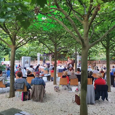 Bild vergrößern: Foto von Menschen, die auf Holzstühlen unter Bäumen sitzen und zur Open-Air-Bühne schauen. Sommerliche Atmosphäre.