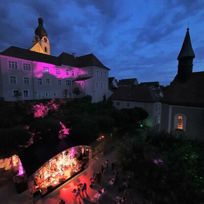 Bild vergrößern: Luftaufnahme der Volkshochschule Schwandorf, die pink angeleuchtet wird. Unterhalb, im Hof des Spitalgartens steht eine Open-Air-Bühne die hellrot erleuchtet ist.