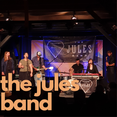 Orangene Schrift auf Foto: "the jules band". Im Hintergrund ist die Musikgruppe auf der Bühne im Sperlstadel des Come Together Festivales 2021.