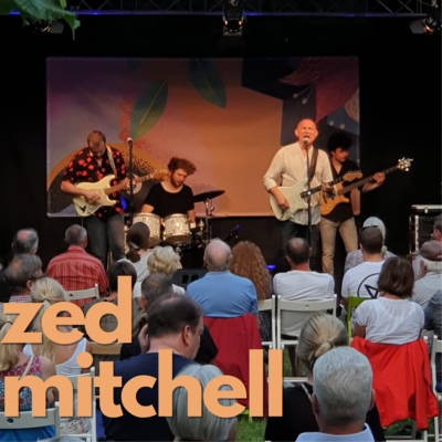 Orangene Schrift auf Foto: "Zed Mitchell"
Im Hintergrund spielt die Musikgruppe um Zed Mitchell auf der Open-Air Bühne des Come Together Festivales 2021.