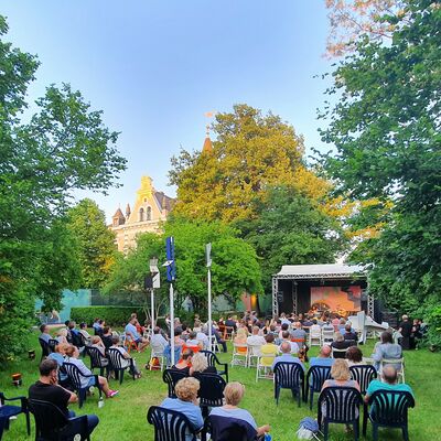 Foto von der Open-air Bühne im Park der Kebbelvilla. Im Vordergrund sitzen die Besucher:innen mit Abstand. Die Sonne scheint.