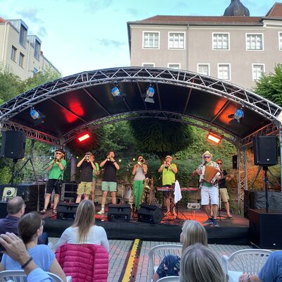 Vier Musikerinnen und Musiker der Band "Skavaria" auf der Bühne des Schwandorfer Kultursommers im Spitalgarten, während sie synchron ein Blasinstrument spielen.