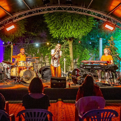 Das Publikum des Schwandorfer Kultursommers im Spitalgarten lauscht den drei Musikern der Band "Tanquoray". Die Open-Air Bühne ist bunt beleuchtet.