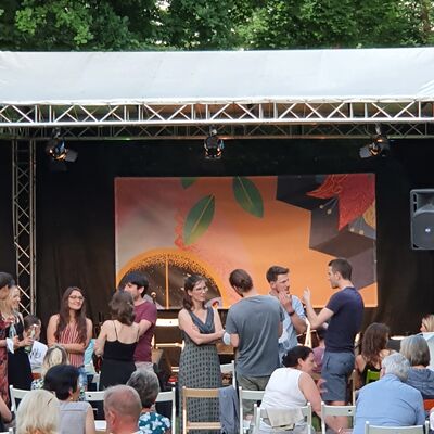 Foto von der Open-Air Bühne, die mit dem Plakat dekoriert ist. Davor stehen Besucher:innen, die miteinander im Gespräch sind.