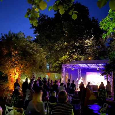 Foto von einem Konzert bei Nacht. Die Bühne ist hell in lila erleuchtet. Die Besucher:innen sitzen oder tanzen an ihren Plätzen.
