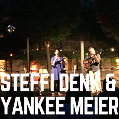 Die Namen der Musiker:innen "Steffi Denk & Yankee Meier" sind in weißen Blockbuchstaben auf dem Foto zu sehen. Im Hintergrund sind die Musiker:innen Steffi Denk und Yankee Meier auf der Open-Air Bühne des Kultursommers 2020 zu sehen.