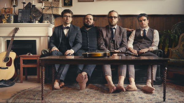 Bild vergrößern: Die vier Musiker der Band Johnny and the Yooahoos sitzen in Anzügen und barfuß an einem niedrigen Wohnzimmertisch.