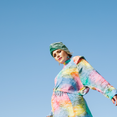 Die K&uuml;nstlerin Die Nowak steht in einem bunten Kleid und mit einer gr&uuml;nen Kopfbedeckung vor einem himmelblauen Hintergrund.