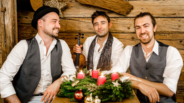 Bild vergrößern: Die drei Musiker von Cobario sitzen in einer Holzhütte um einen Adventskranz, bei dem alle vier Kerzen brennen.