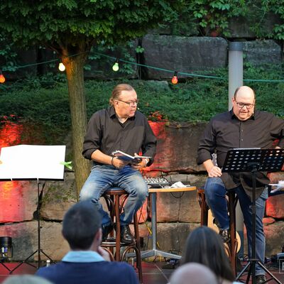 Foto: Die Künstler "Schmarrnkerl & Funkerl" tragen, sitzend auf der Open-Air-Bühne, ihr Programm mündlich aus blauen Heften vor.