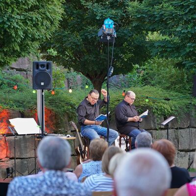 Foto: Die Künstler "Schmarrnkerl & Funkerl" tragen, sitzend auf der Open-Air-Bühne, ihr Programm mündlich aus blauen Heften vor.