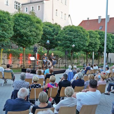 Foto: Die Künstler "Schmarrnkerl & Funkerl" tragen sitzend auf der Open-Air-Bühne ihr Programm mündlich aus blauen Heften vor. Davor sitzen Besucher:innen, die auf die Bühne schauen.