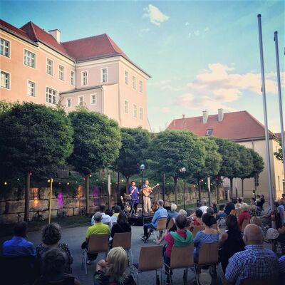 Foto der Open-Air Location im Spitalgarten. Zahlreiche Personen sitzen vor der Bühne. Es ist Sommerabend.