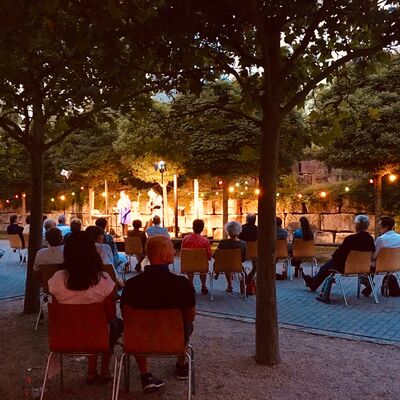 Foto: Besucher:innen sitzen im nächtlichen Spitalgarten vor der hell-erleuchteten Open-Air-Bühne. Künstler:innen performen auf der Bühne.