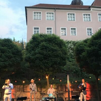 Foto: Auf einer erleuchteten Open-Air Bühne spielen 4 Musiker der Musikgruppe Bavaschoro beim Kultursommer im Spitalgarten 2020. Sommerabendliche Stimmung.