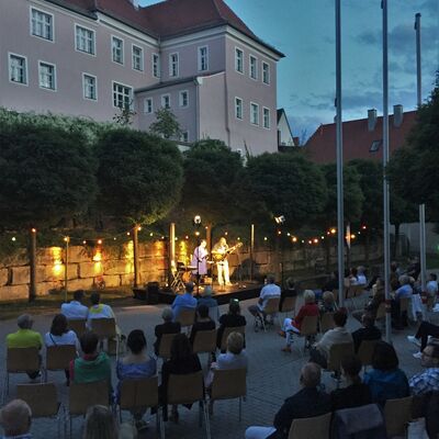 Foto: Die hell erleuchtete Open-Air-Bühne nach dem Sonnenuntergang. Vor dieser sitzen Menschen auf Stühlen. Die Bäume sind erleuchtet. Auf der Bühne sind die Musiker:innen Steffi Denk und Yankee Meier zu sehen.