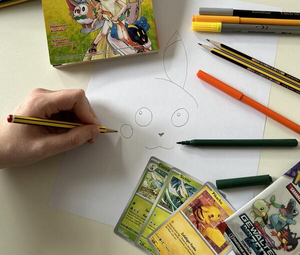Ein Hand zeichnet eine Pokemonfigur, um die Zeichnung herum liegen Pokemon Karten.