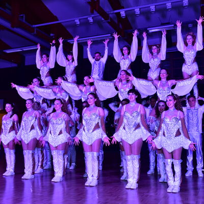 Aufnahme einer akrobatischen Tanzchoreografie. Die Gruppe ist mit weißen Bühnenoutfits gekleidet. Teilweise stehen sie auf den Schultern der Anderen.